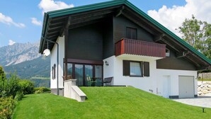Una casa moderna de dos pisos amb teulada verda, un balcó i vista a la muntanya.