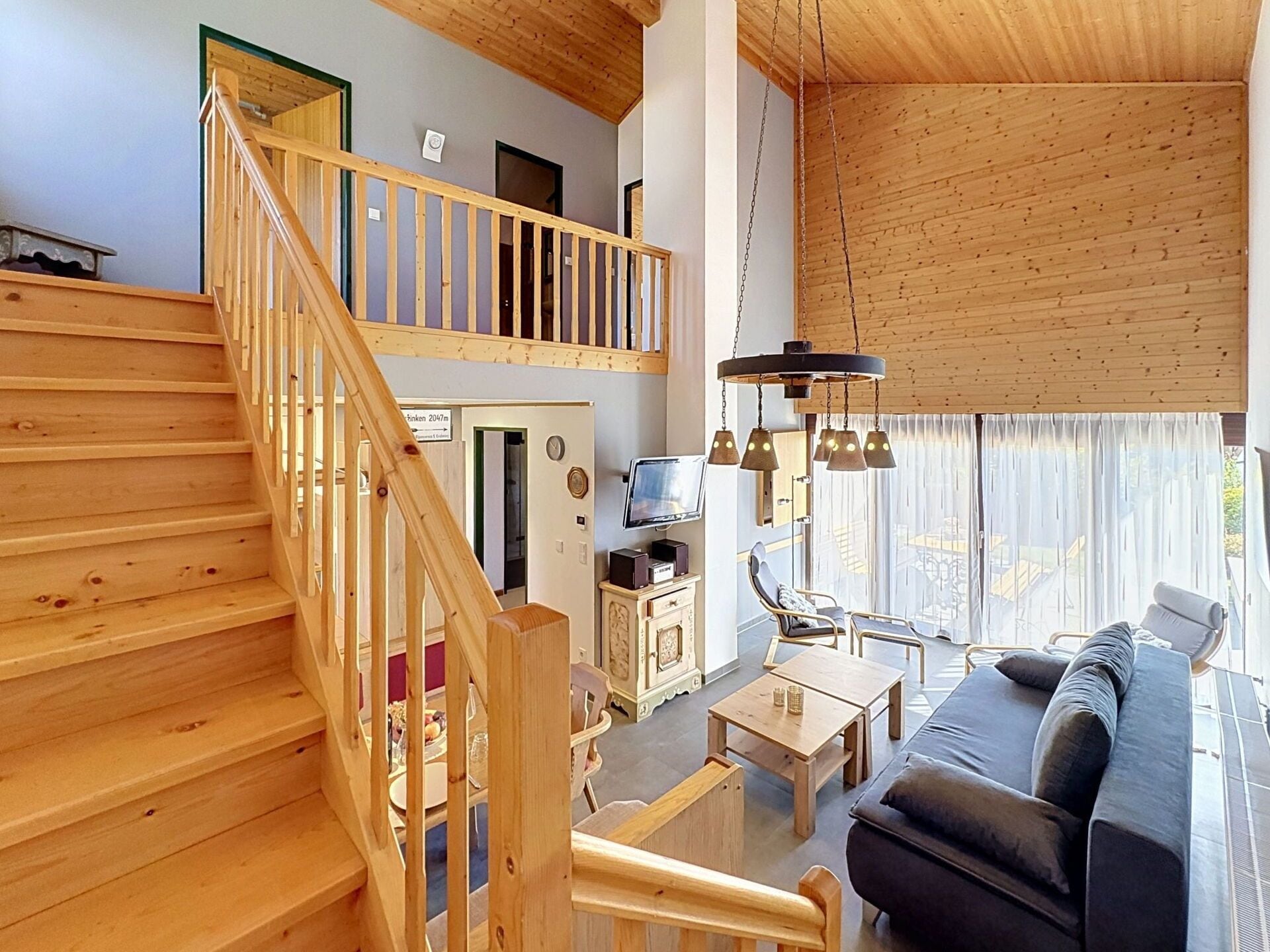 Un escalier en bois mène à une chambre située en mezzanine, équipée d’une télévision et d’un coin repas avec une table et des chaises.