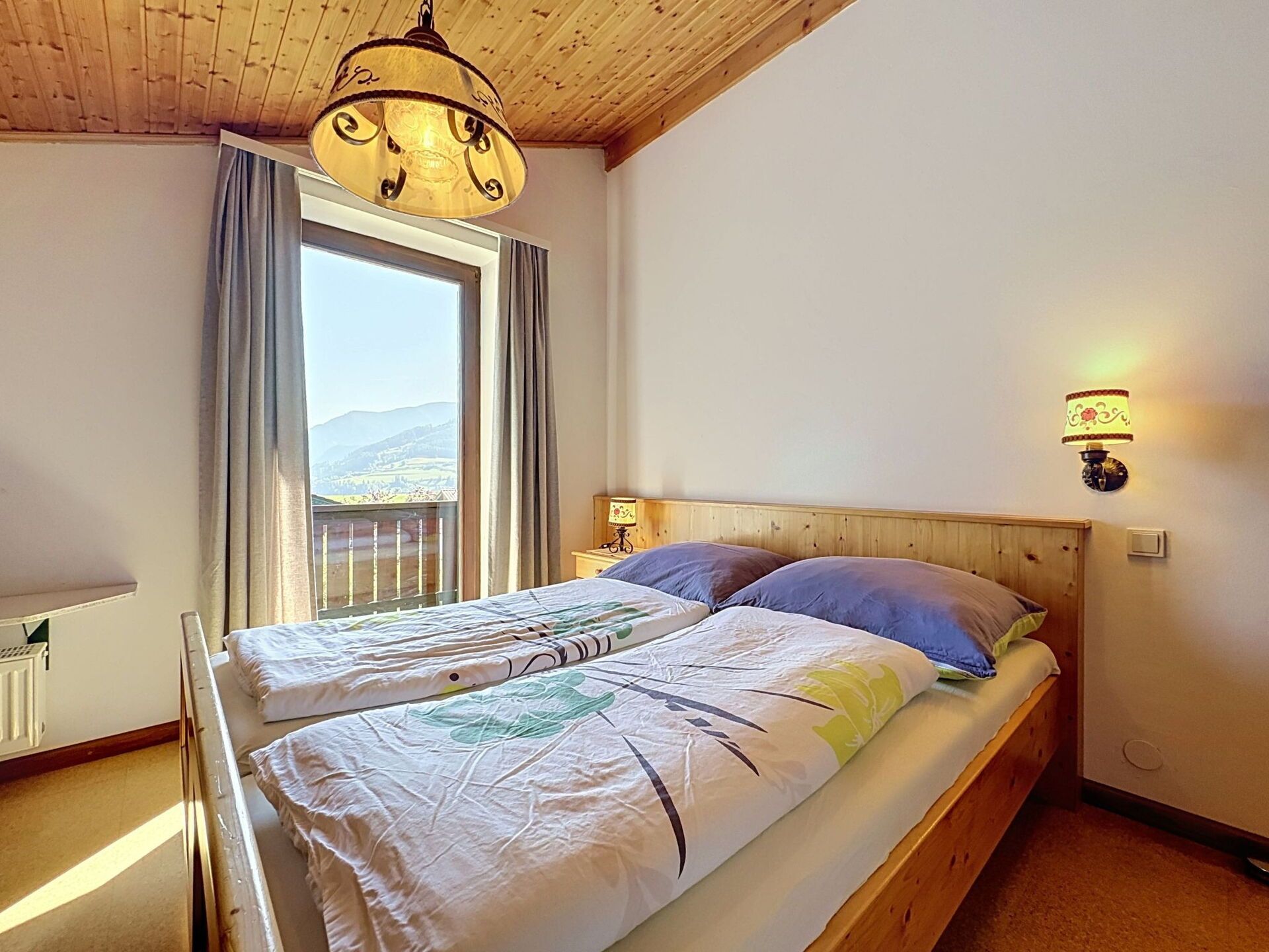 Een slaapkamer met een houten hoofdbord, een bed met twee kussens, een raam met gordijnen en een wandlamp.