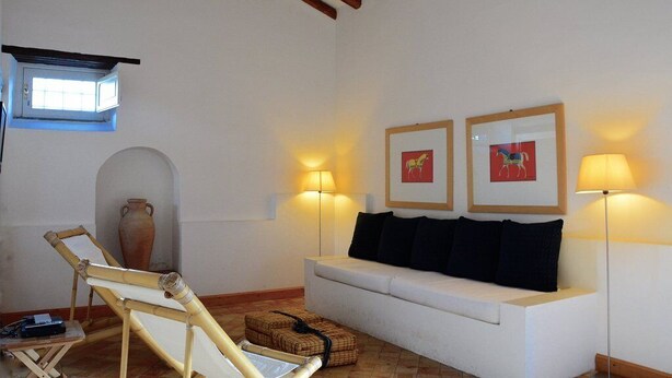 Une pièce avec un canapé blanc, deux lampes fixées au mur, un tableau encadré, une chaise en bois et une petite table.