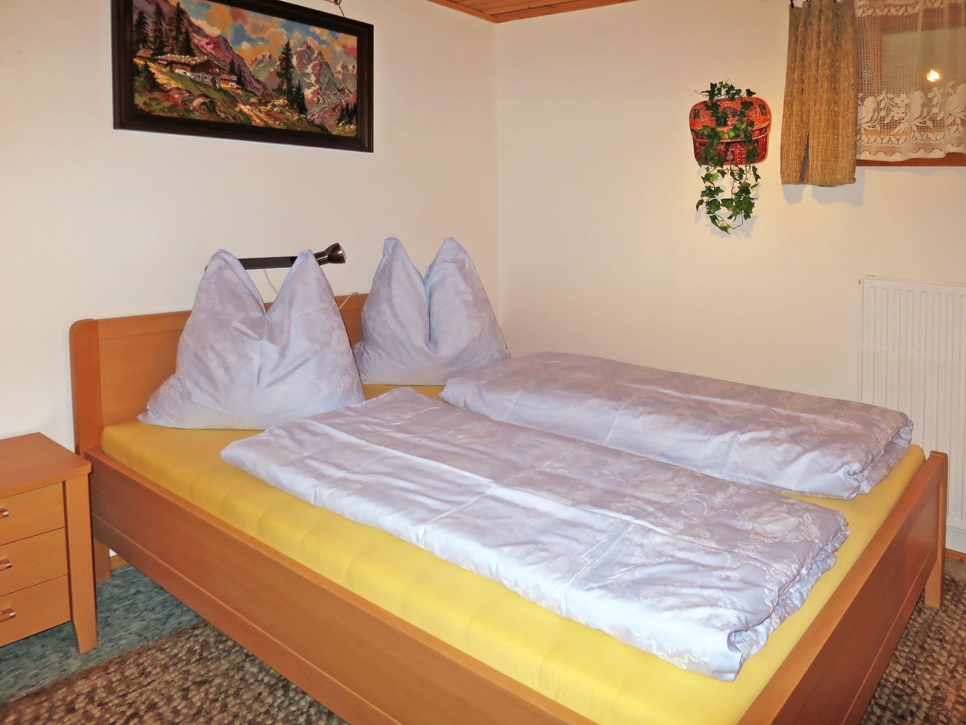 Ein Schlafzimmer mit einem Doppelbett, zwei Kissen, einem Nachttisch, einem Heizkörper, einer Wandpflanze und einem Gemälde.