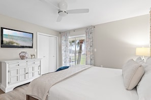 Une chambre à coucher avec un lit, un téléviseur fixé au mur, un ventilateur de plafond, une fenêtre avec des rideaux et une table de chevet avec une lampe.