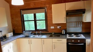 A kitchen with wooden cabinets, a stove, a sink, and a window with a view of greenery. - Goldcrest 3 Hot Tub (Carmarthen)