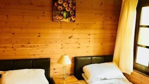 A room with wooden walls, two beds with white linens, a nightstand with a lamp, and a window with curtains. - Goldcrest 3 Hot Tub (Carmarthen)