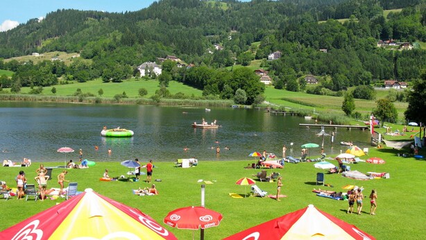 A lakeside resort with grassy areas, umbrellas, and people enjoying the water. - Cosy apartment for 4 guests with WIFI, TV, patio and pets allowed (Dietrichstein)