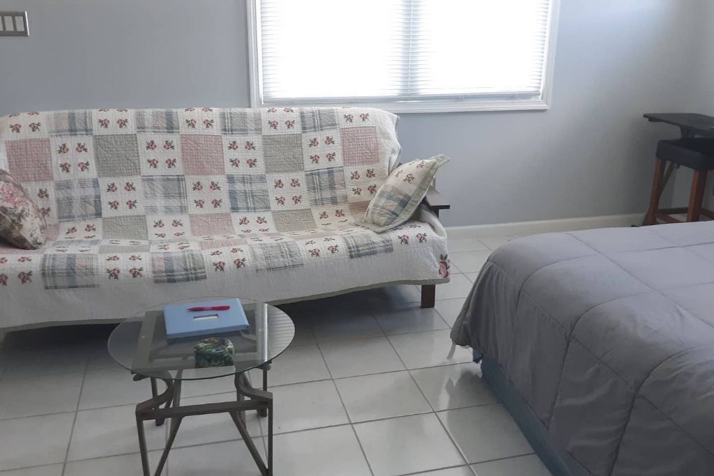 Une pièce avec un canapé recouvert d’une courtepointe à motifs floraux, une petite table ronde sur laquelle se trouve un classeur bleu, et un lit avec une couette grise.