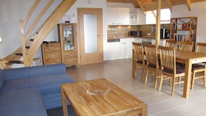 Ein gemütliches Wohnzimmer mit einer Couch, einem Holzespeisetisch und Stühlen. Im Hintergrund ist eine Küche mit weißen Schränken und einer Mikrowelle erkennbar.
