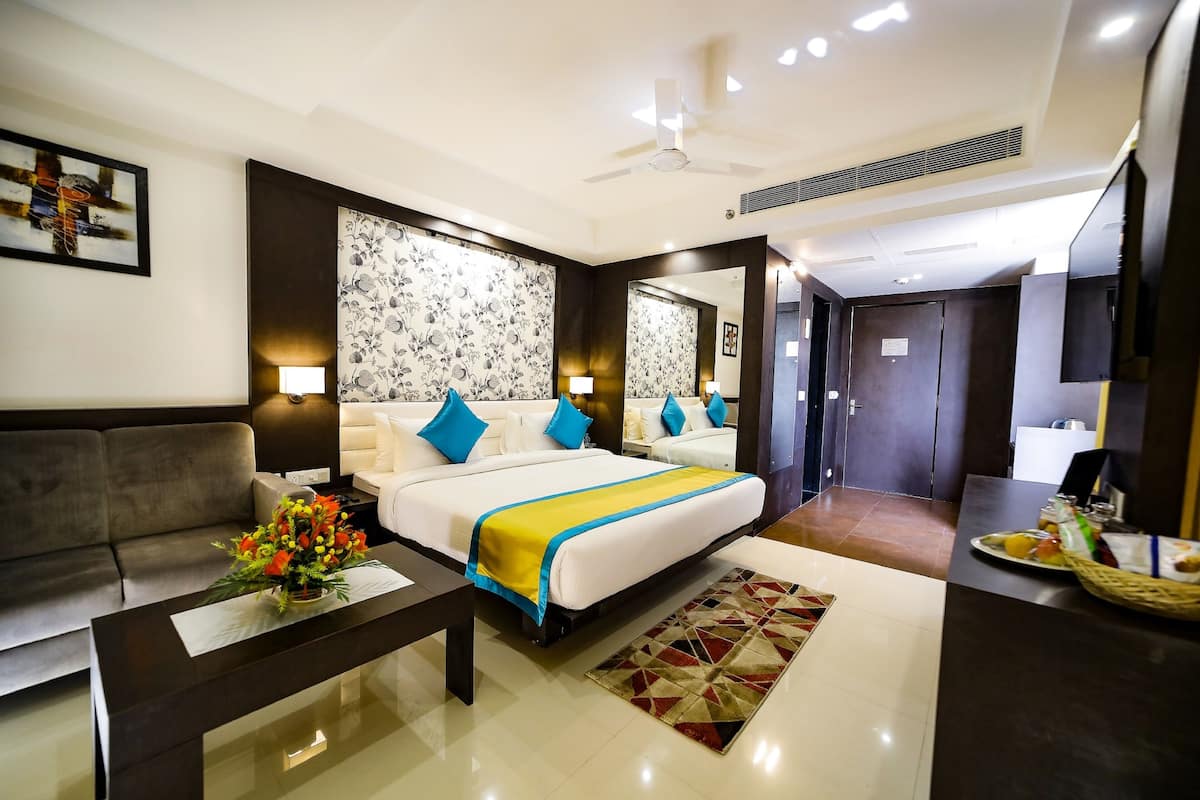 Habitación de hotel con una cama grande, un sofá, una mesa de centro con flores, una mesa de comedor con comida y un espejo.