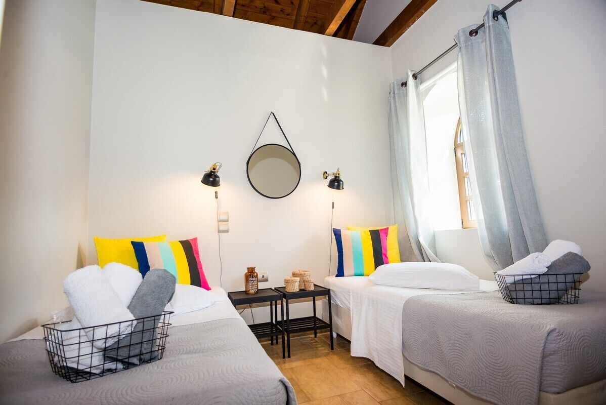 A hotel room with two beds, a small table with baskets, a mirror on the wall, and a window with curtains.