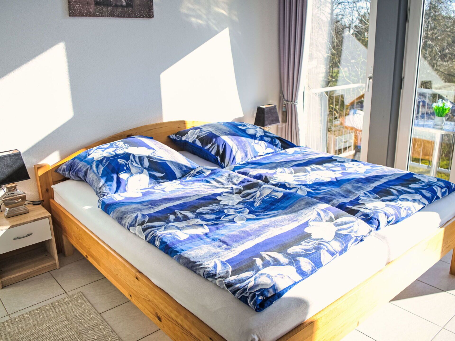 A bed with a floral bedspread, a bedside table with a lamp, and a view of the outdoors through a window.