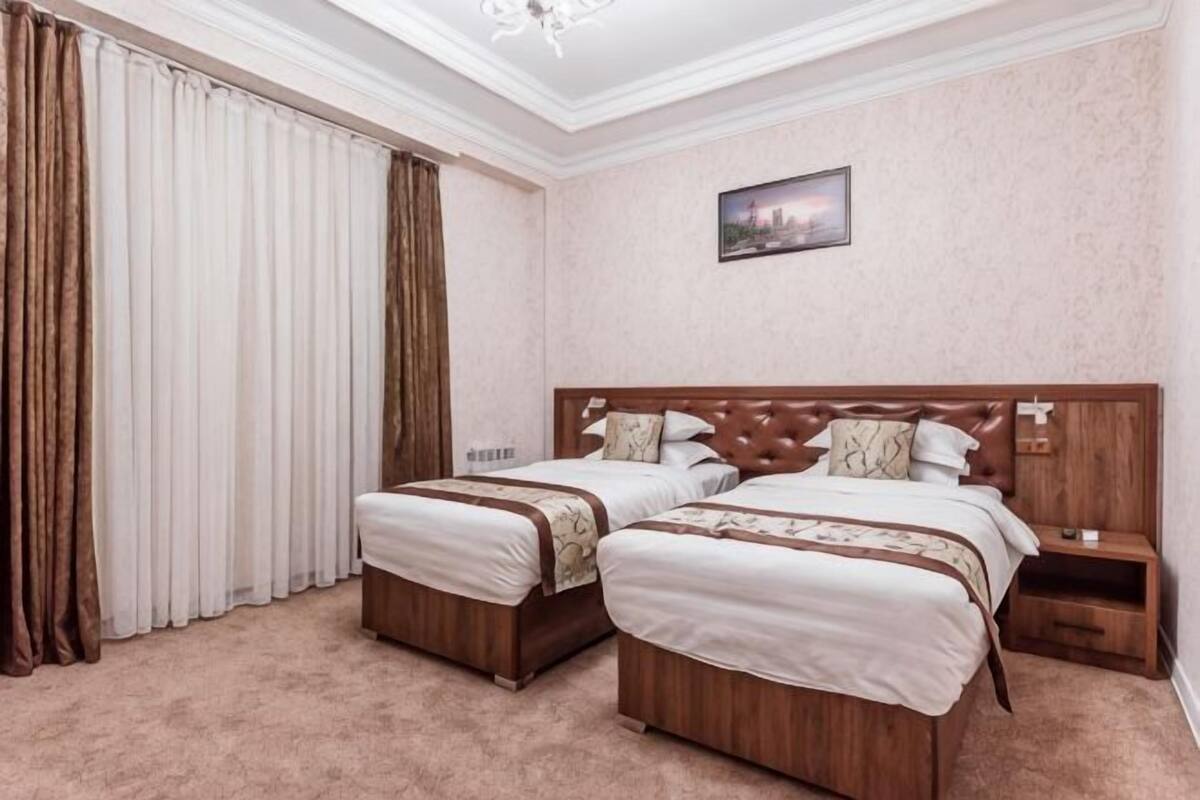 A double room with two beds, a nightstand, a wooden headboard, a window with curtains, and a painting on the wall.