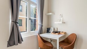 Una habitación pequeña y luminosa con un escritorio blanco, dos sillas marrones, un ventanal con cortinas grises y una estantería blanca con una botella de vidrio y una cafetera.