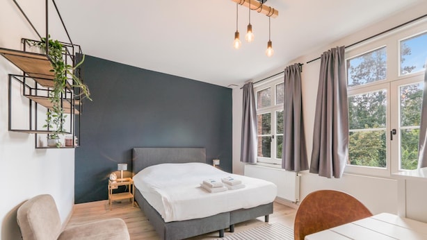 Un dormitorio moderno con cama, mesita de noche, lámpara y vistas al verdor exterior.