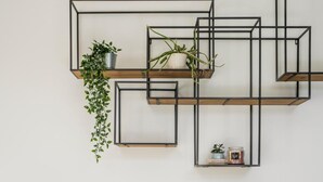 Una habitación moderna con una cama, una silla marrón, una pequeña mesa redonda y un estante en la pared con plantas y una vela.