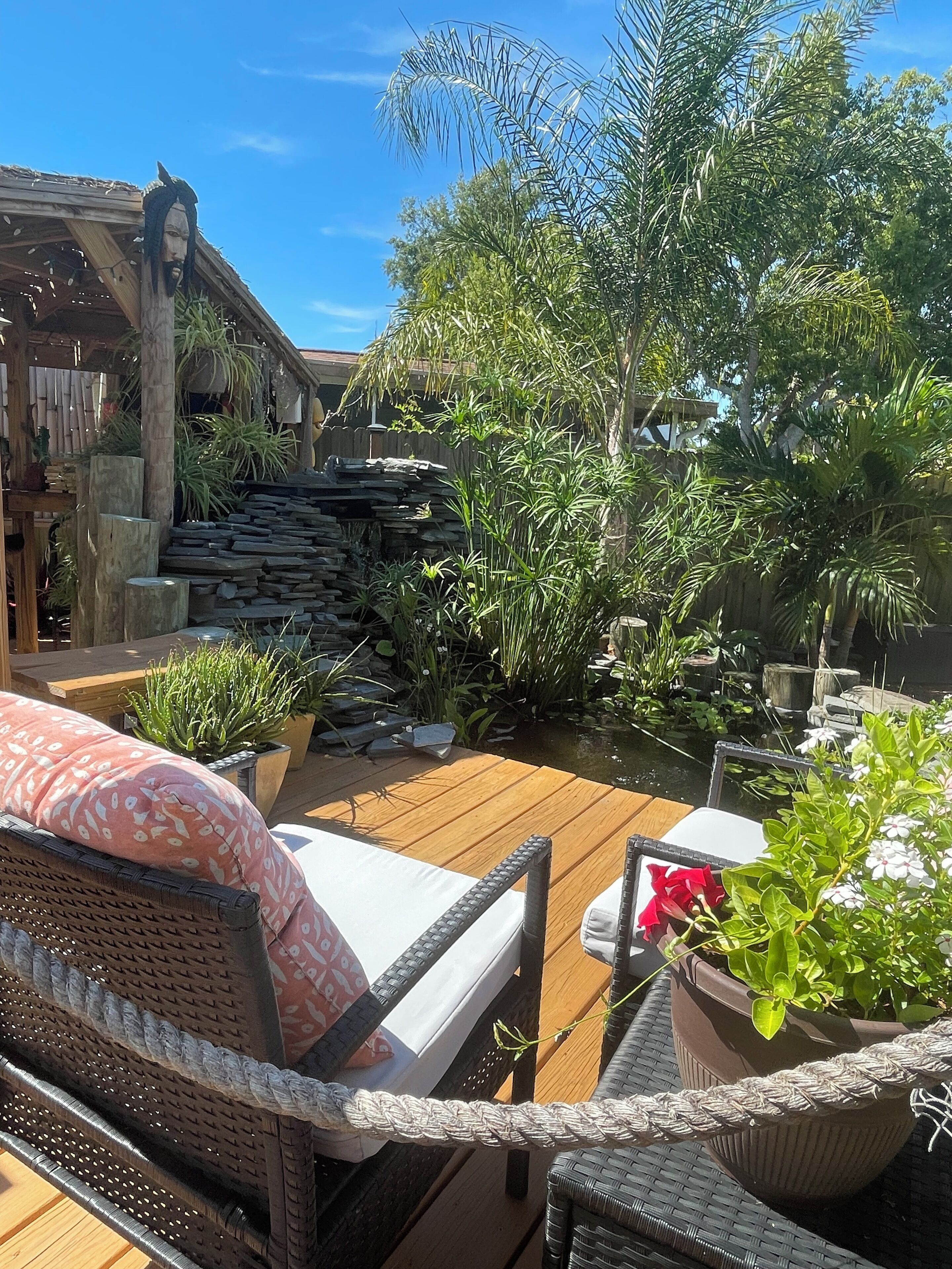 A wooden deck with wicker furniture, surrounded by lush greenery and a stone wall.