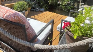 A wooden deck with wicker furniture, surrounded by lush greenery and a stone wall. - Waterfront home w/dock-Jon boat, Kayaks & fishing poles included! (Port Richey)