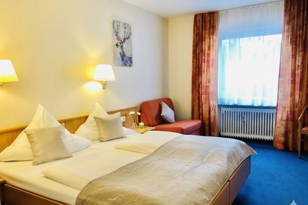 A hotel room with a large bed, two bedside lamps, a red chair, a window with curtains, and a radiator.