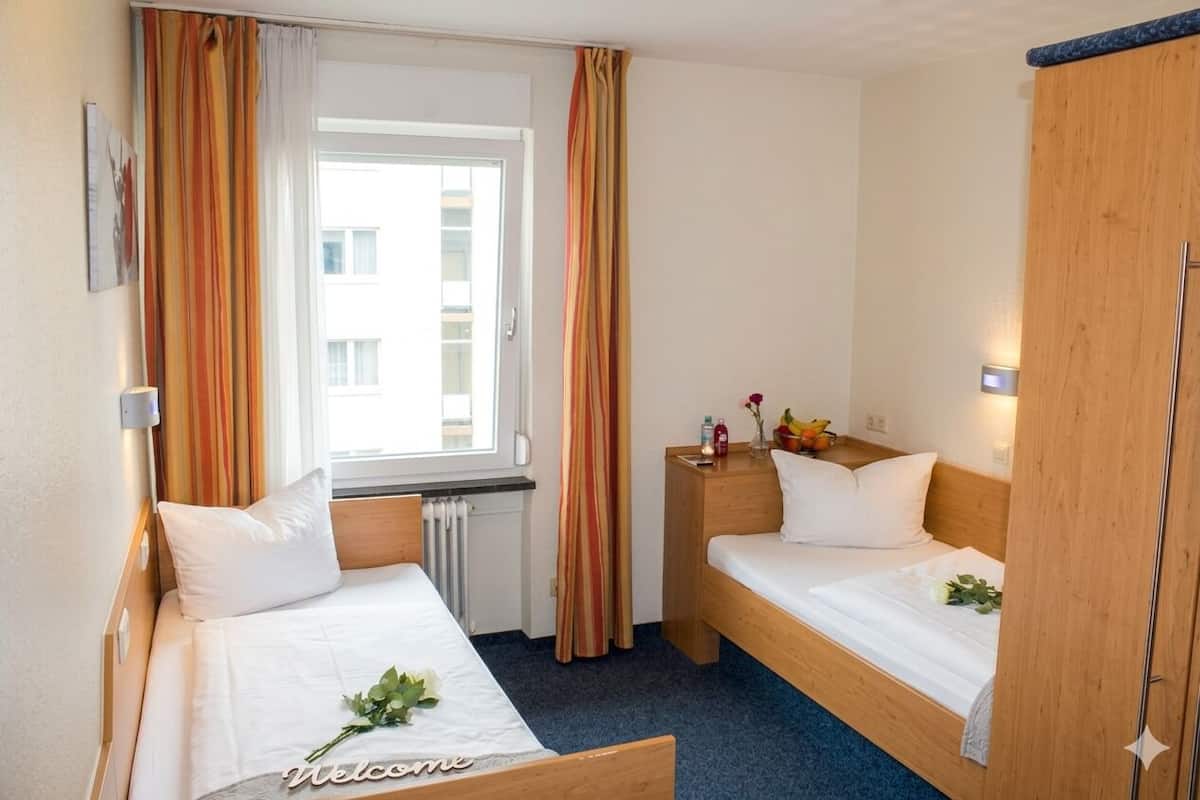 A hotel room with two single beds, a window with curtains, a bedside table with a lamp, and a wooden wardrobe.