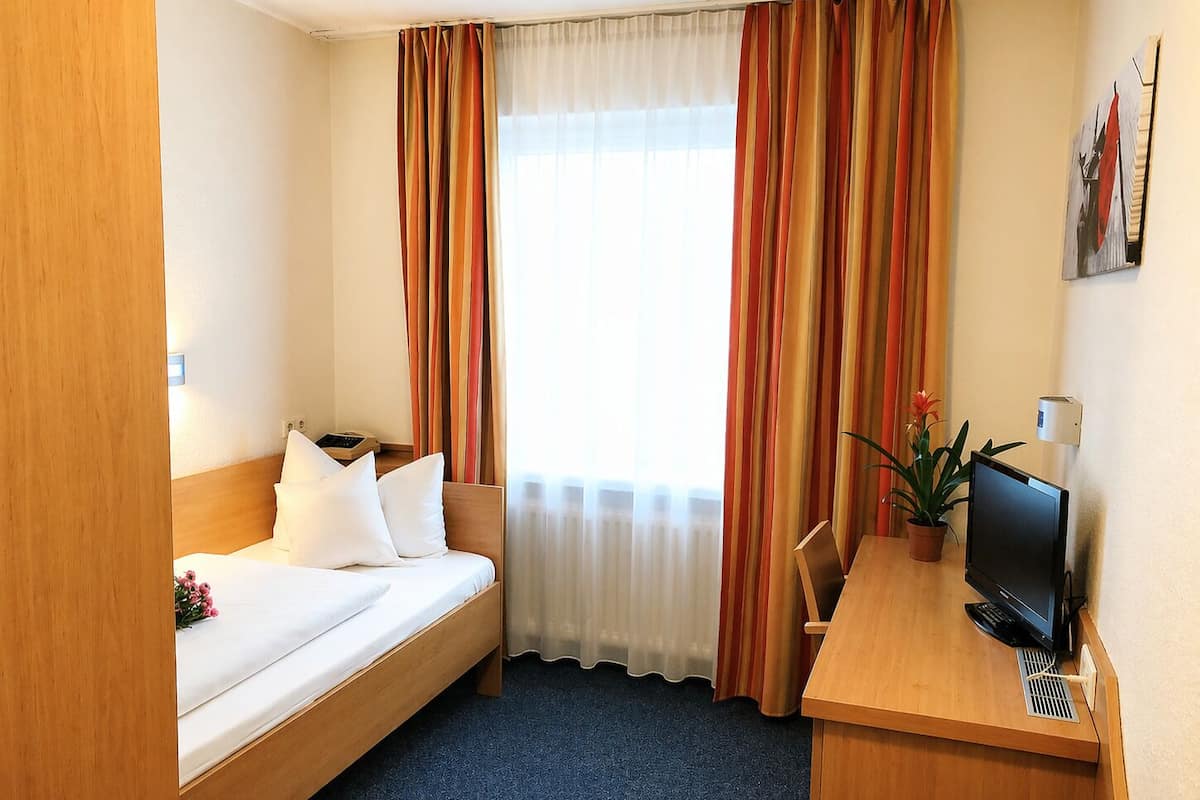 A hotel room with a bed, a desk with a computer, a window with curtains, and a potted plant.