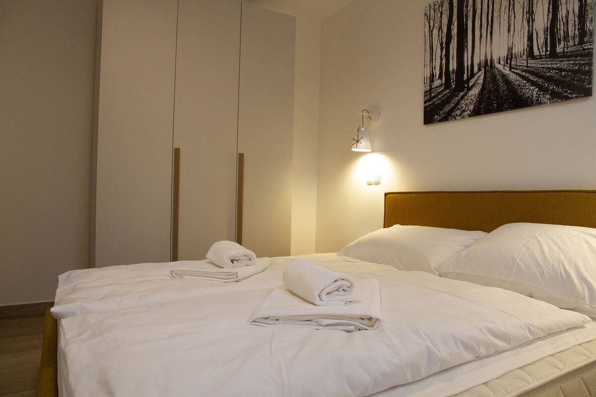 Un lit bien fait, avec du linge de lit et des serviettes blancs, une tête de lit en bois et un luminaire fixé au mur.