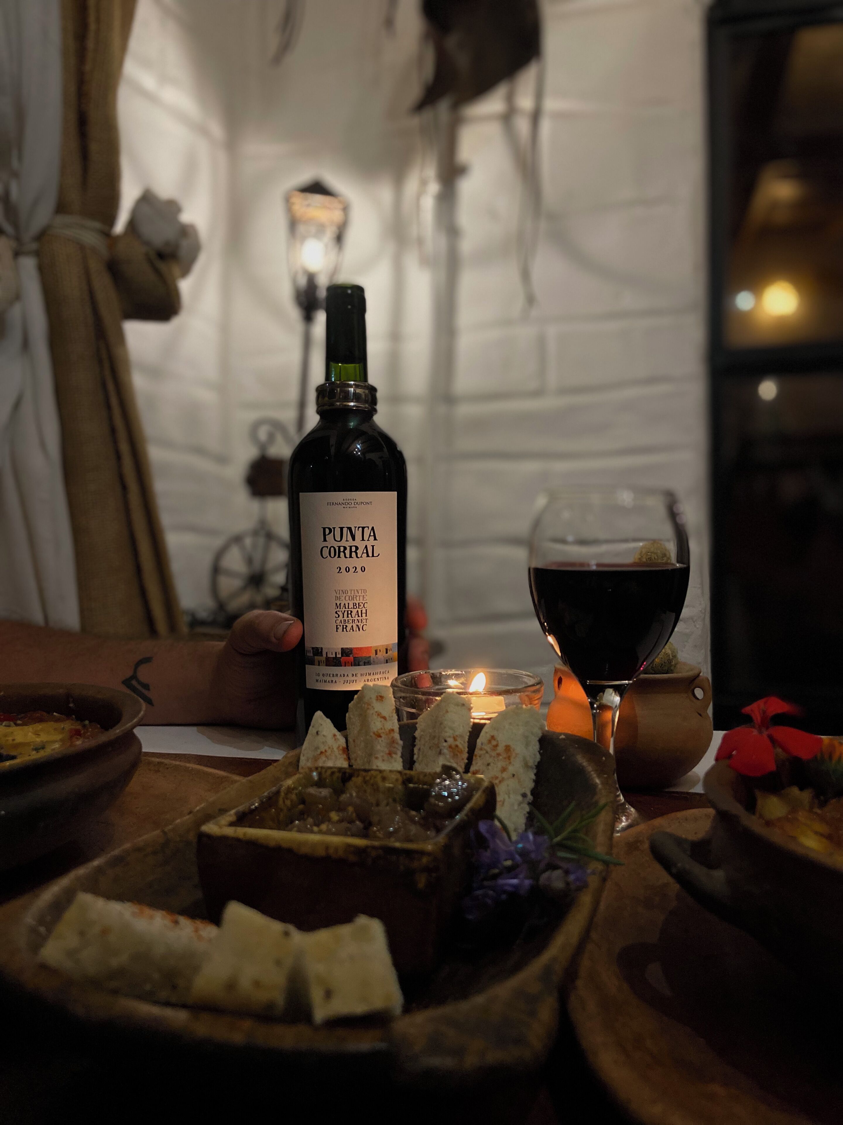 A bottle of Punta Corral 2020 wine, a glass of red wine, and a candle on a table with food.