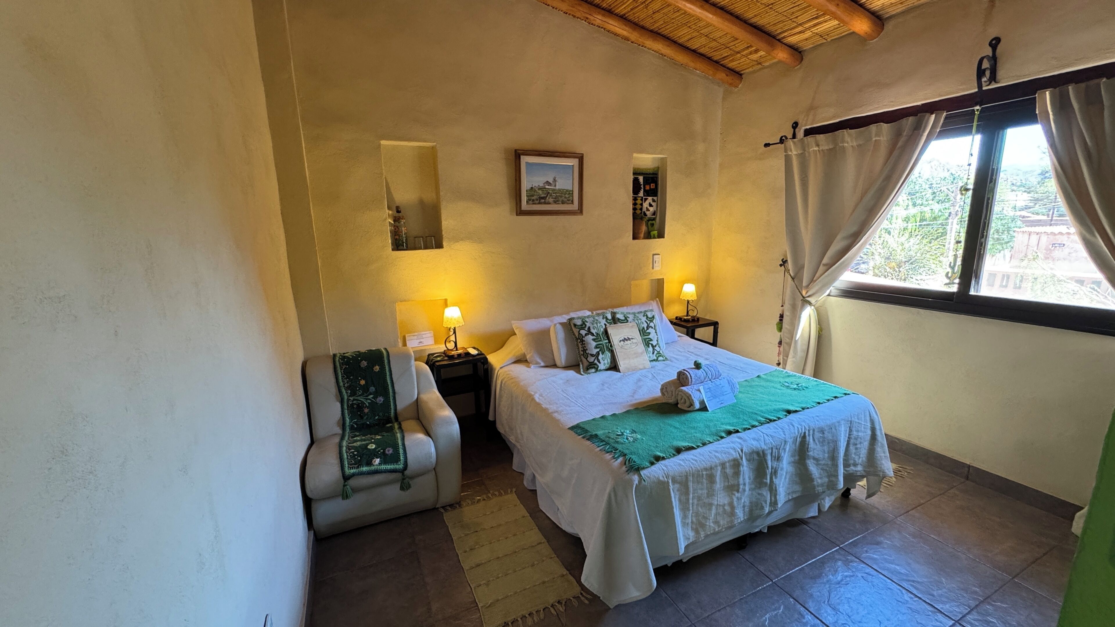 Un dormitorio con una cama, dos lámparas de noche, una silla, una mesita, un espejo y una ventana con cortinas.