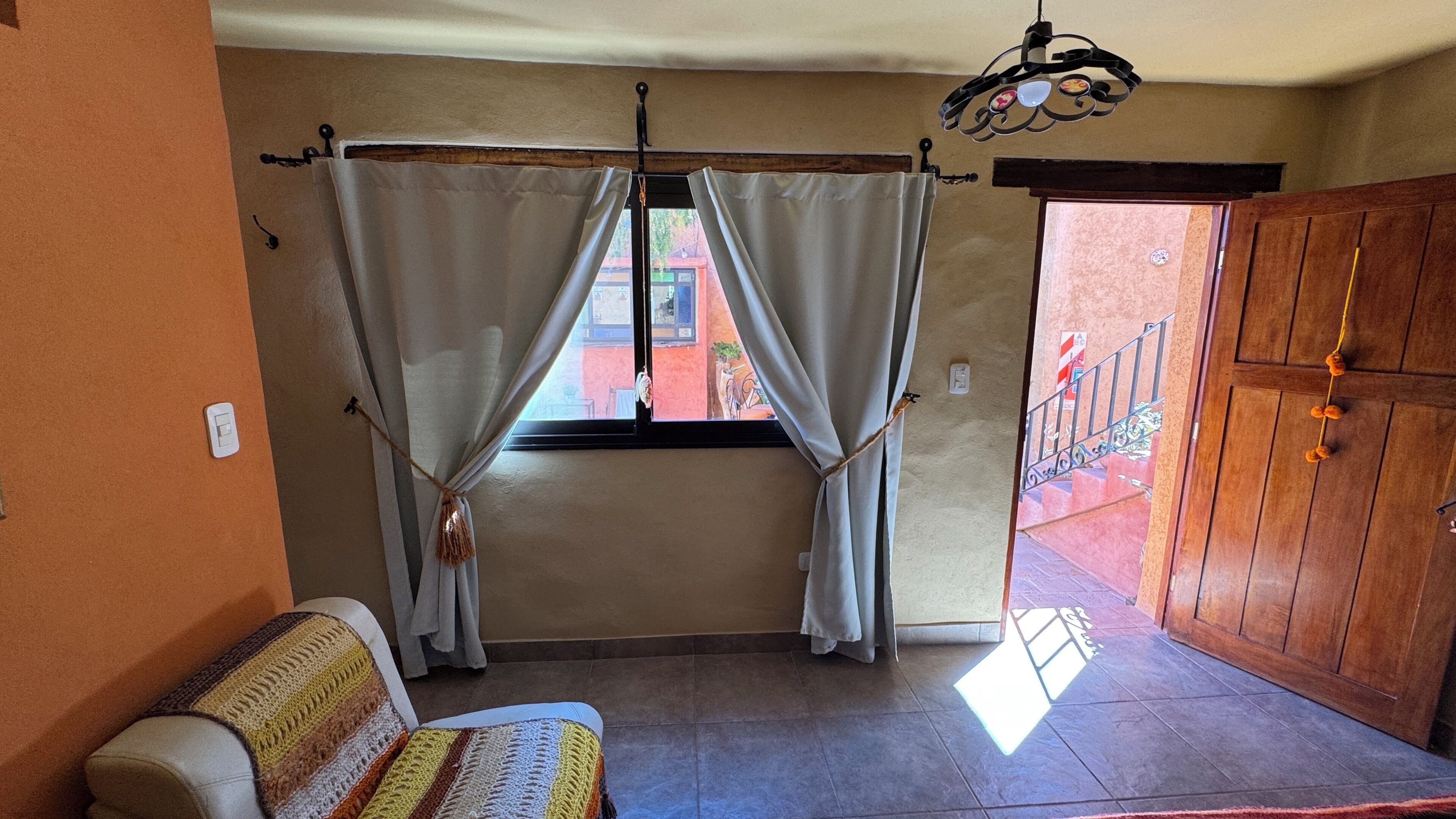 Una habitació amb un llit, una finestra amb cortines, una làmpada penjant i una porta de fusta.