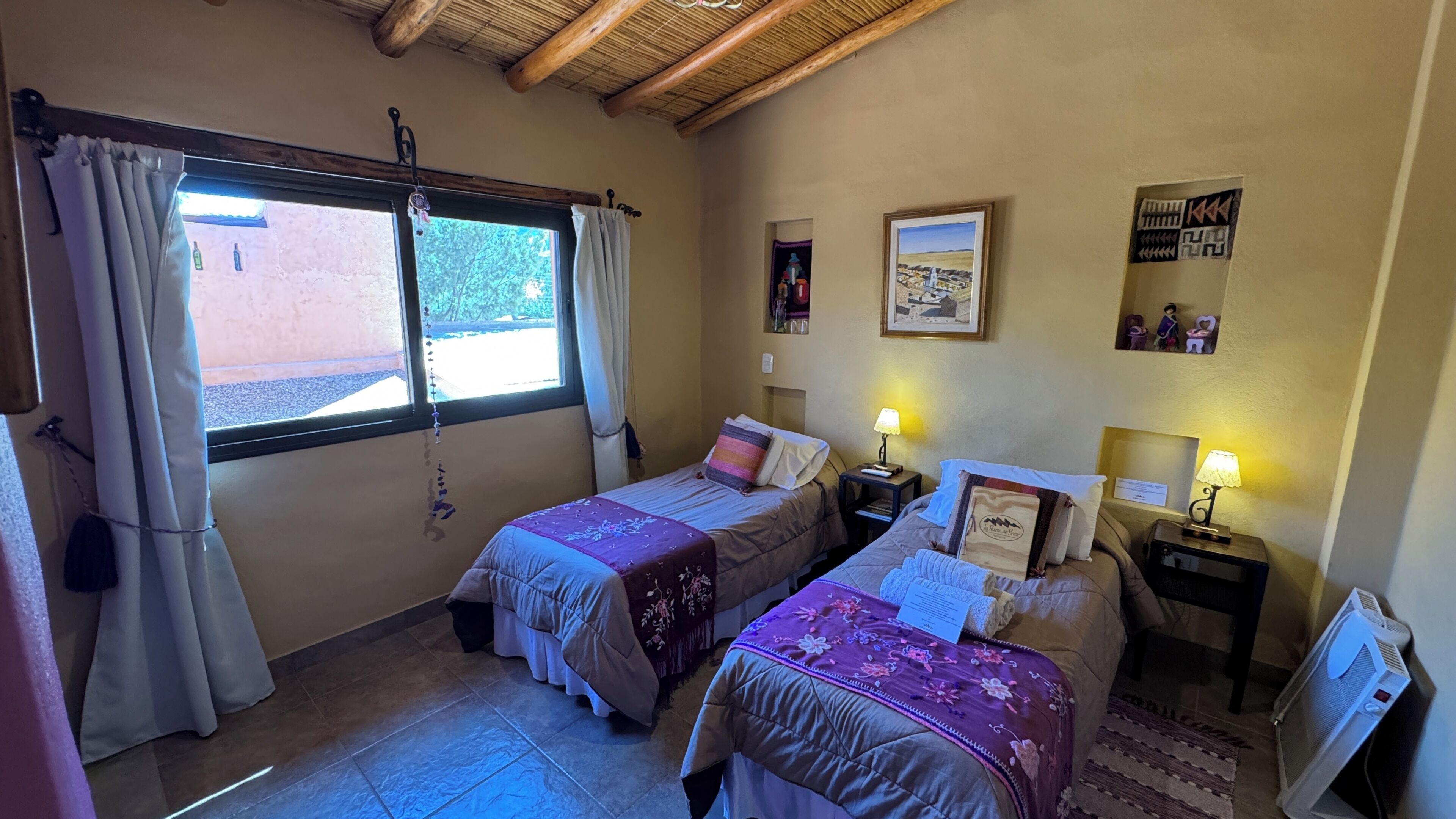 Une chambre avec deux lits, une fenêtre avec des rideaux, un mur orné de tableaux encadrés et un radiateur.