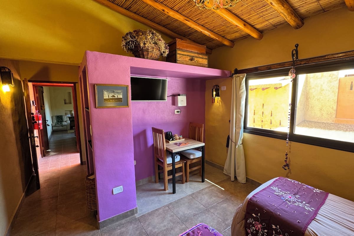 Une pièce avec un mur violet, un plafond en bois, une fenêtre avec des rideaux, une table avec des chaises et un lit recouvert d’une couverture violette.