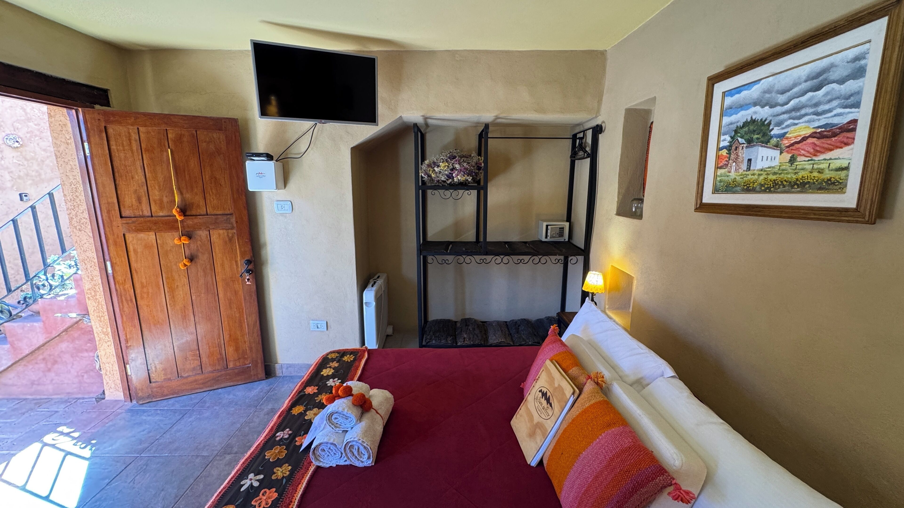 Una habitación con una cama, una puerta de madera, un televisor montado en la pared y un cuadro.