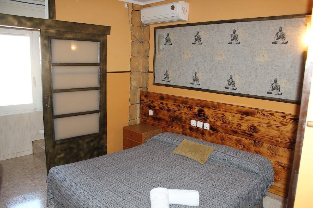 A bedroom with a bed, a wooden headboard, a wall-mounted air conditioner, and a framed picture of meditating figures.