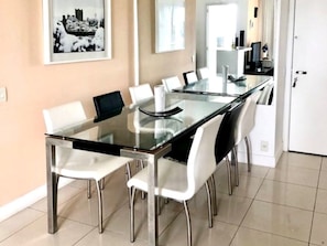 A modern dining area with a glass table and white chairs, a kitchen in the background, and a door on the right.