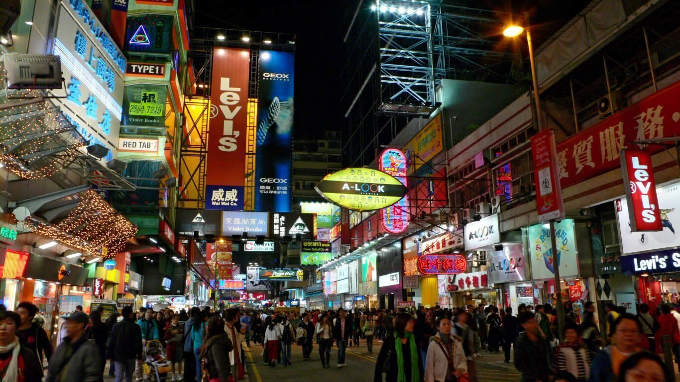 Una calle urbana concurrida, iluminada por carteles y vallas publicitarias, incluyendo marcas como Levi's y A-LOOK.