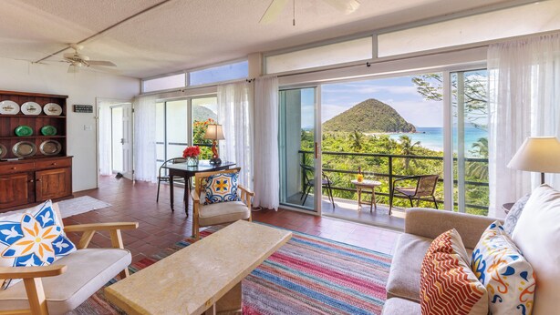 A spacious living area with a balcony overlooking a scenic ocean view, a ceiling fan, and a dining area with a table and chairs. - This stunning View Could Be Yours (West End)