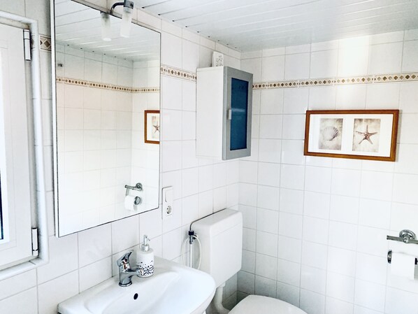 Un baño con paredes de azulejos blancos, un lavamanos con dispensador de jabón, un inodoro y un espejo. Hay cuadros enmarcados en la pared.