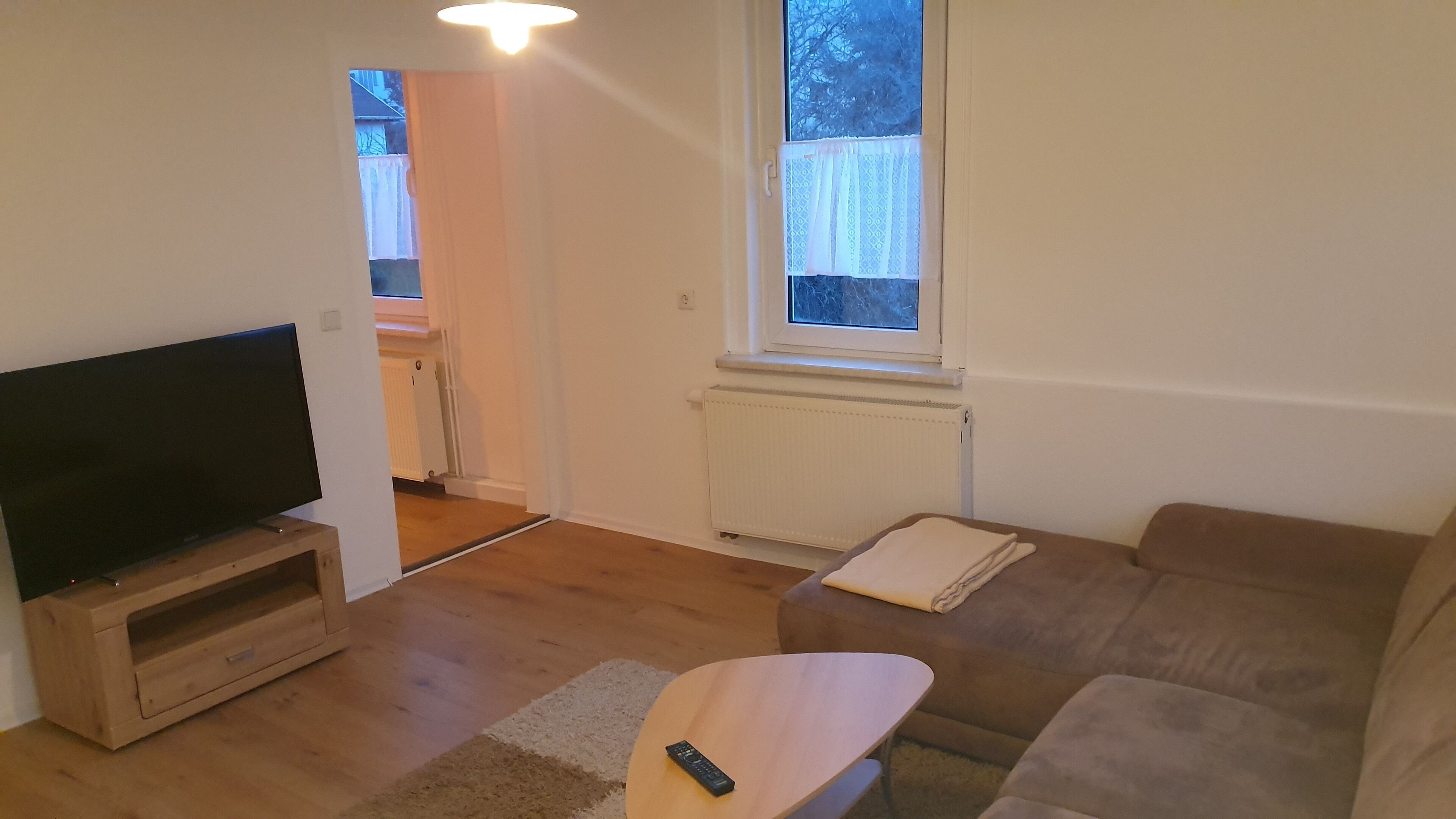 A living room with a flat-screen TV, a wooden TV stand, a brown sofa, a small wooden table, and a remote control on the table.