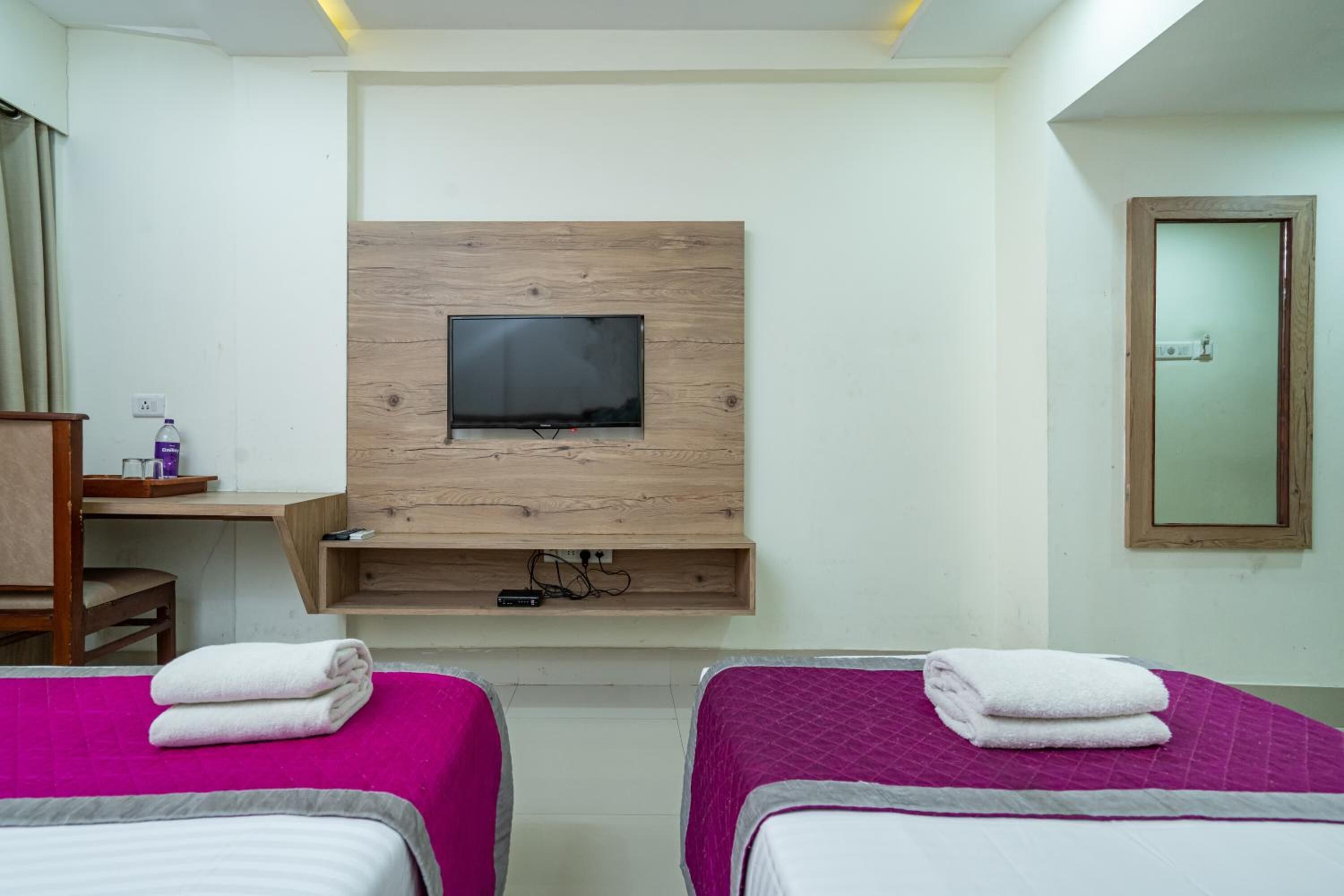 A hotel room with two beds, a TV mounted on a wooden panel, a desk with a chair, and a mirror on the wall.