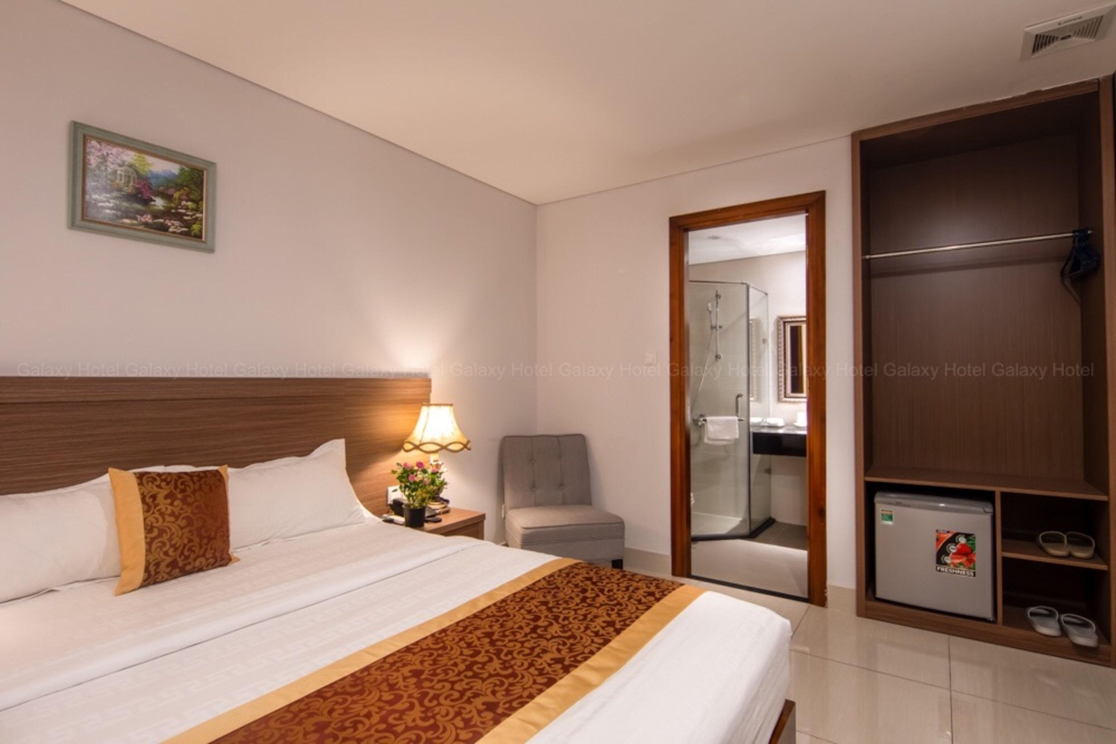 A hotel room with a large bed, a nightstand with a lamp, a chair, a wardrobe, and a small refrigerator.