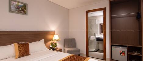 A hotel room with a large bed, a nightstand with a lamp, a chair, a wardrobe, and a small refrigerator.