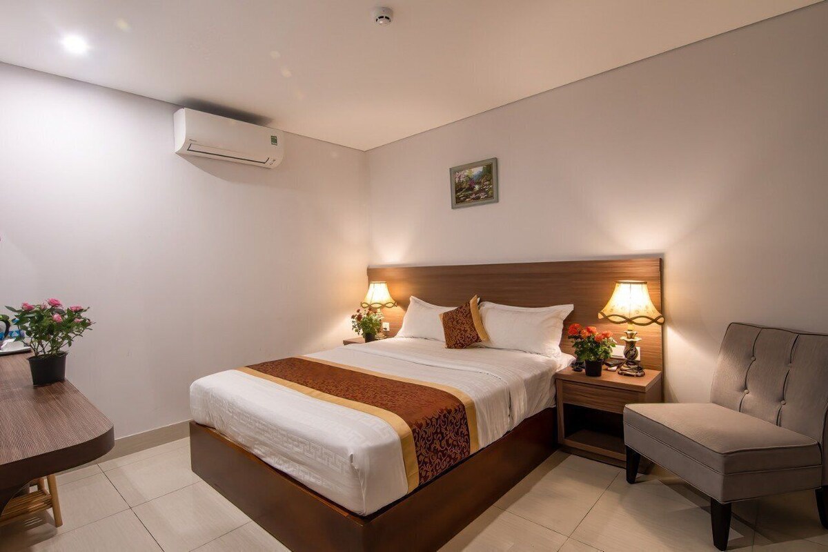 A hotel room with a bed, bedside lamps, a chair, a small table with a plant, and an air conditioning unit.