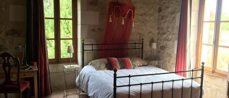 Un dormitorio rústico con estructura de cama metálica, dosel, escritorio con silla y ventana con cortinas.