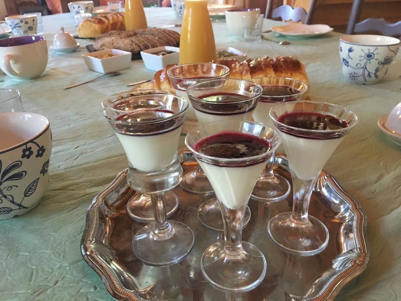 Uma mesa posta com taças de sobremesa repletas de creme branco e compota de frutas vermelhas, um croissant e uma garrafa de suco de laranja.