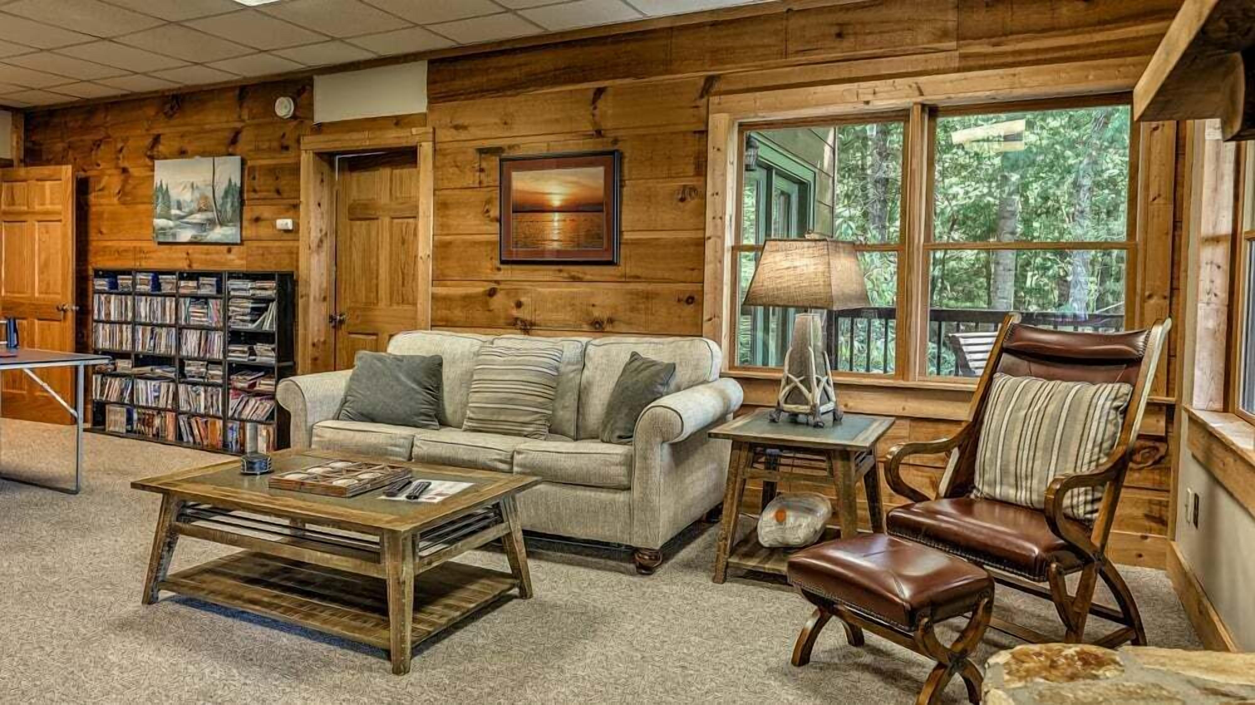 A cozy cabin interior with a wooden interior, a sofa, a coffee table, a leather armchair, and a bookshelf filled with books.