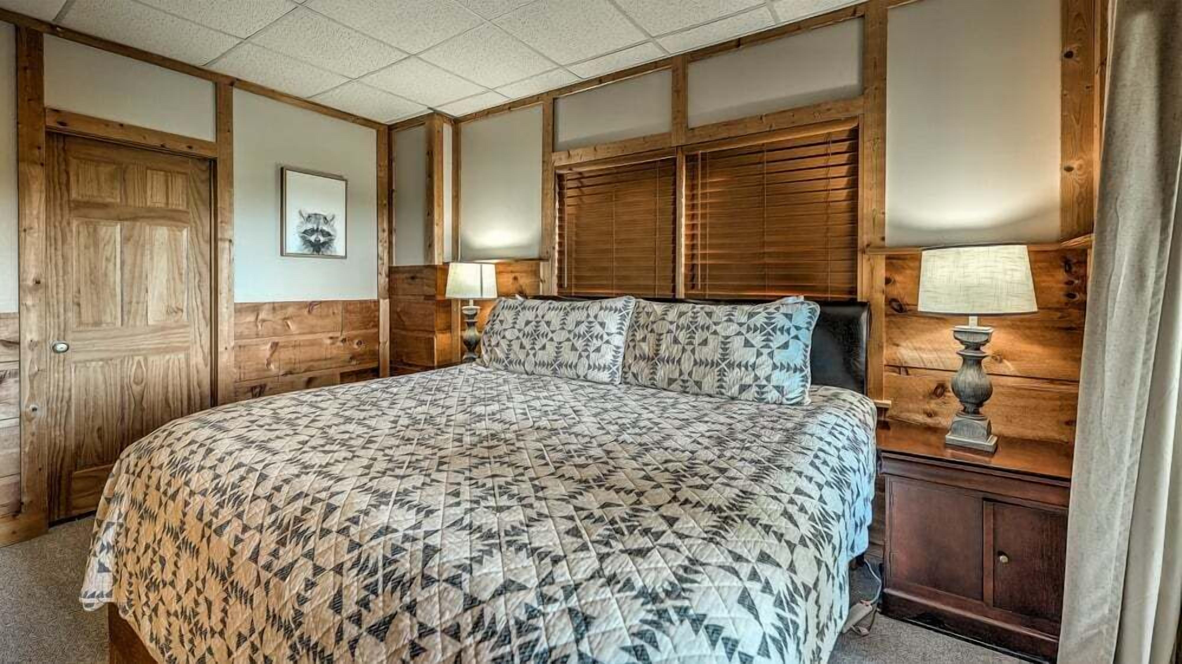 A bedroom with a large bed, wooden walls, a wooden door, and a nightstand with a lamp.