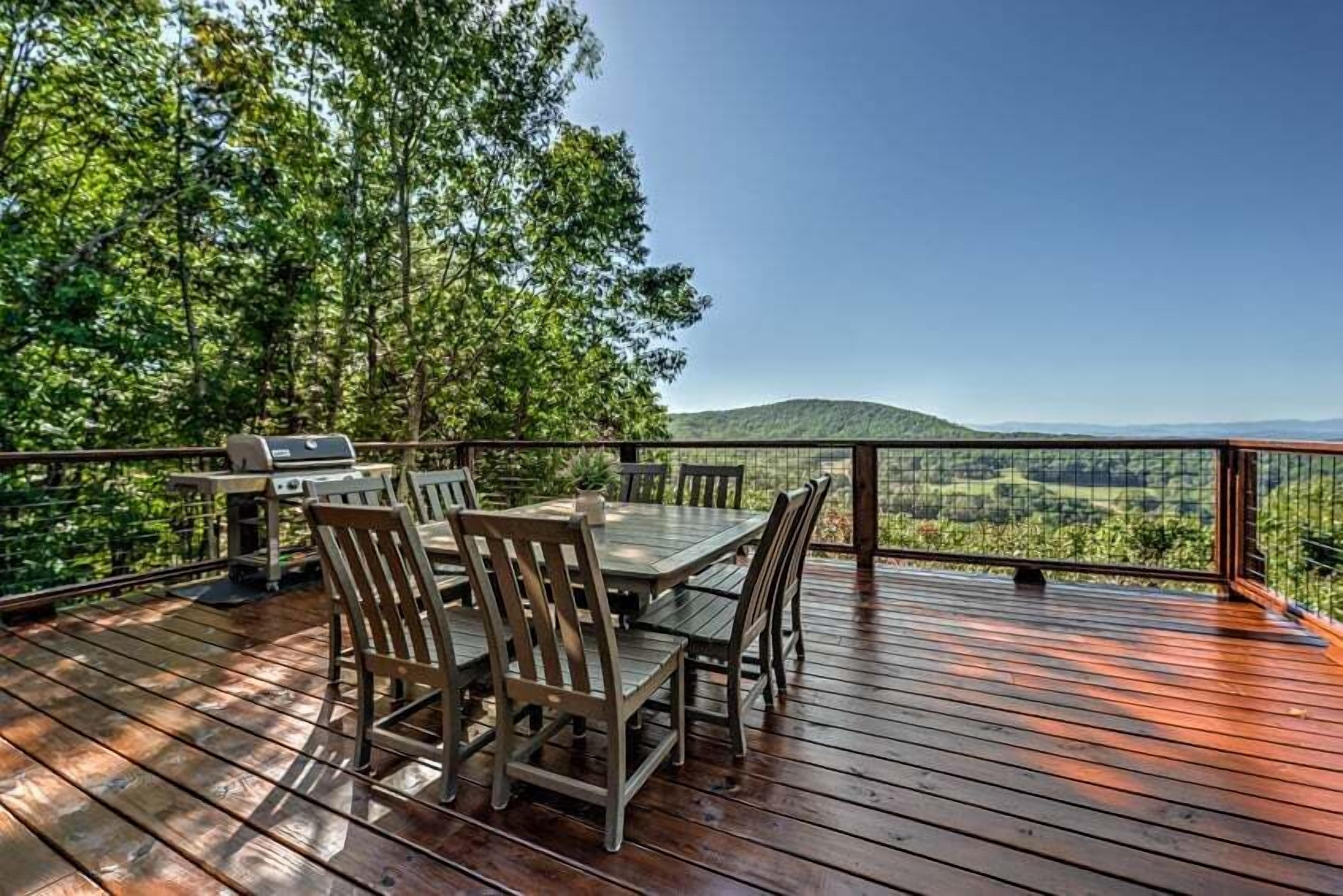 A wooden deck with a table and chairs, overlooking a forested area and distant hills.