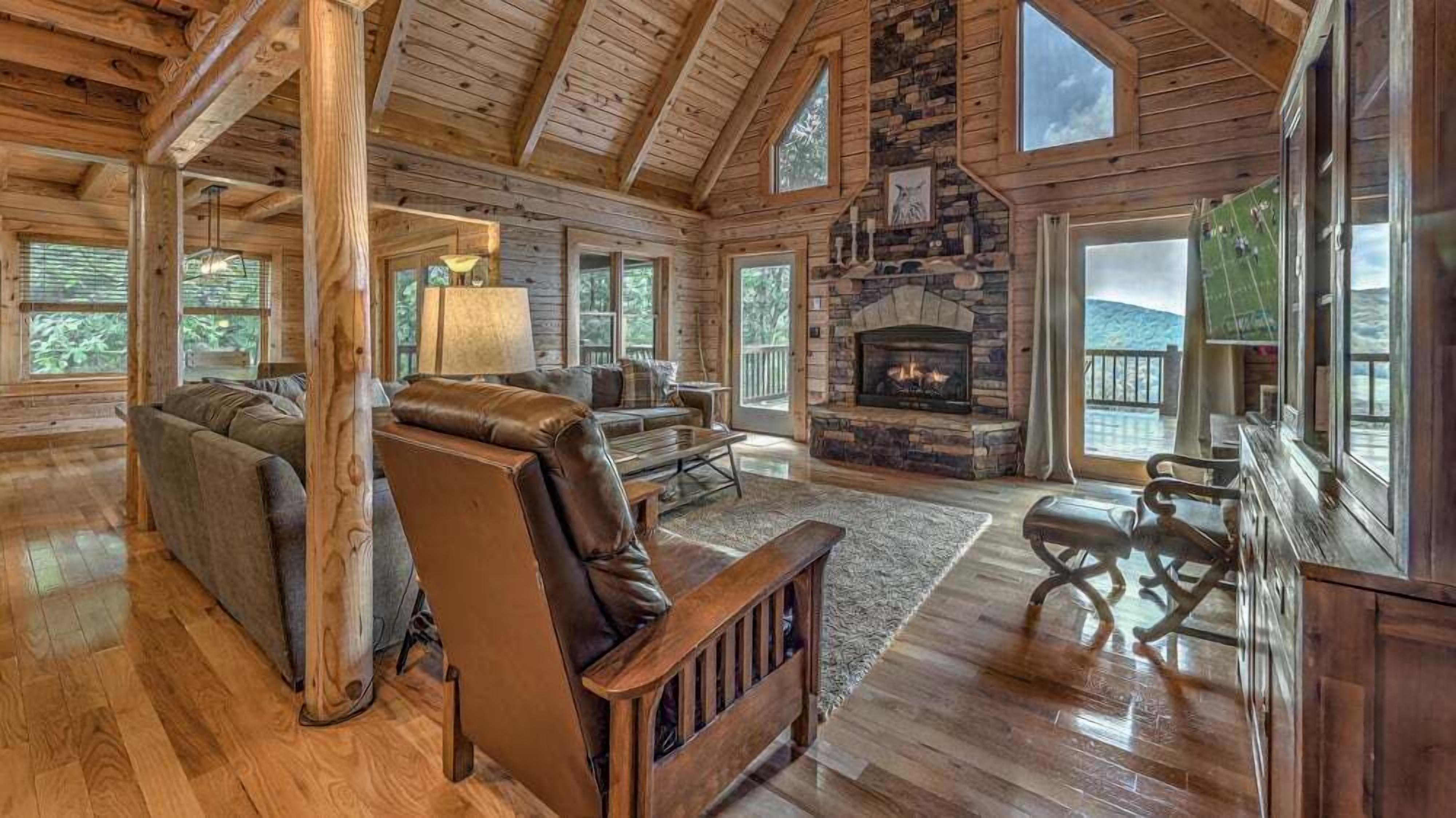 Un salón espacioso con chimenea de piedra, vigas de madera y grandes ventanales que ofrecen vistas al exterior.