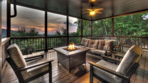 A covered outdoor seating area with a fire feature, wooden furniture, and a view of a forested landscape.
