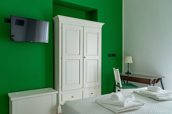 Una camera d'albergo con una parete verde, un armadio bianco, un letto con lenzuola bianche, una scrivania con una lampada e un televisore montato a parete.