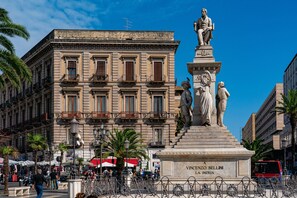 Una piazza pubblica con una statua di un uomo e una donna, panchine e palme.
