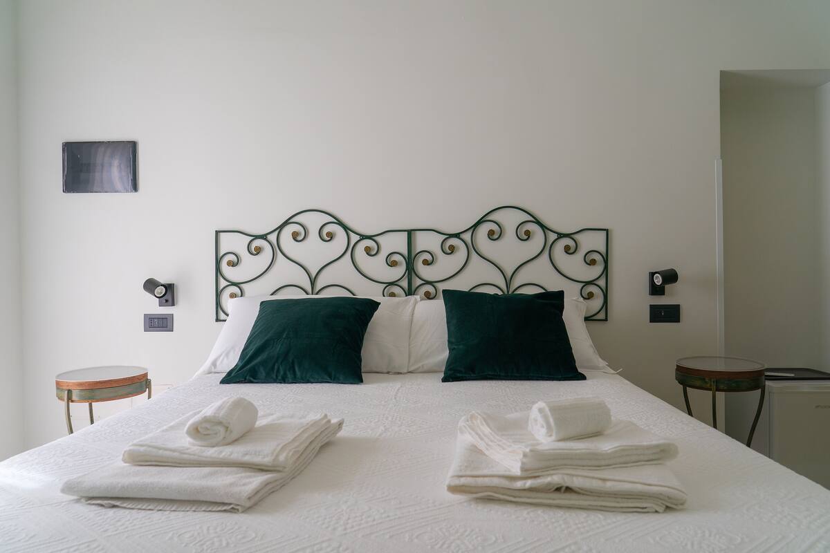 Un letto rifatto con lenzuola bianche e due cuscini verdi, un piccolo tavolino rotondo e un lampadario fissato alla parete.